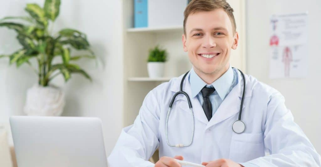 A smiling doctor wearing a white lab coat and stethoscope sitting at a desk with a laptop.