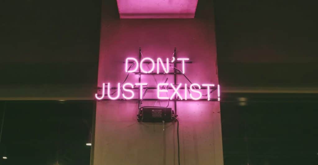 A neon pink sign on a dark wall with the words "DON'T JUST EXIST!".