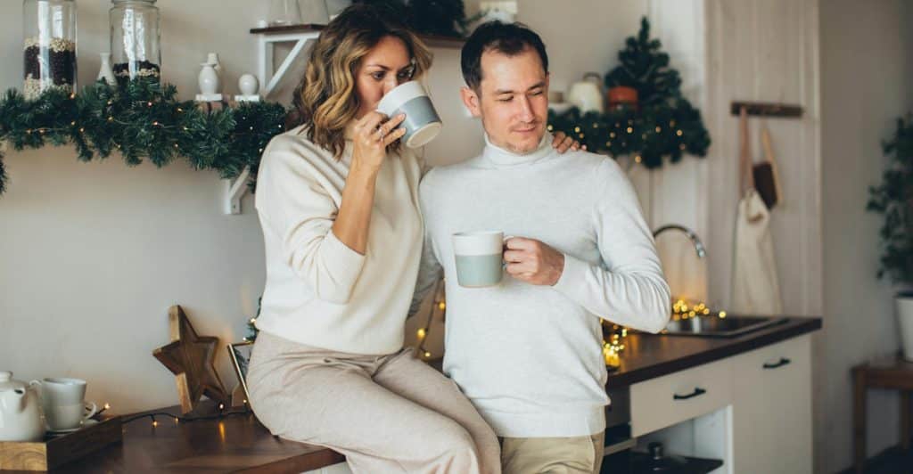 A man and a woman in cozy sweaters holding mugs and standing close in a kitchen decorated with festive greenery.