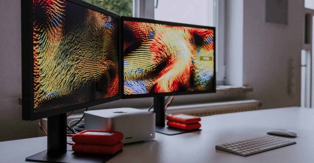Two computer monitors displaying colorful abstract images on a desk with a keyboard and mouse