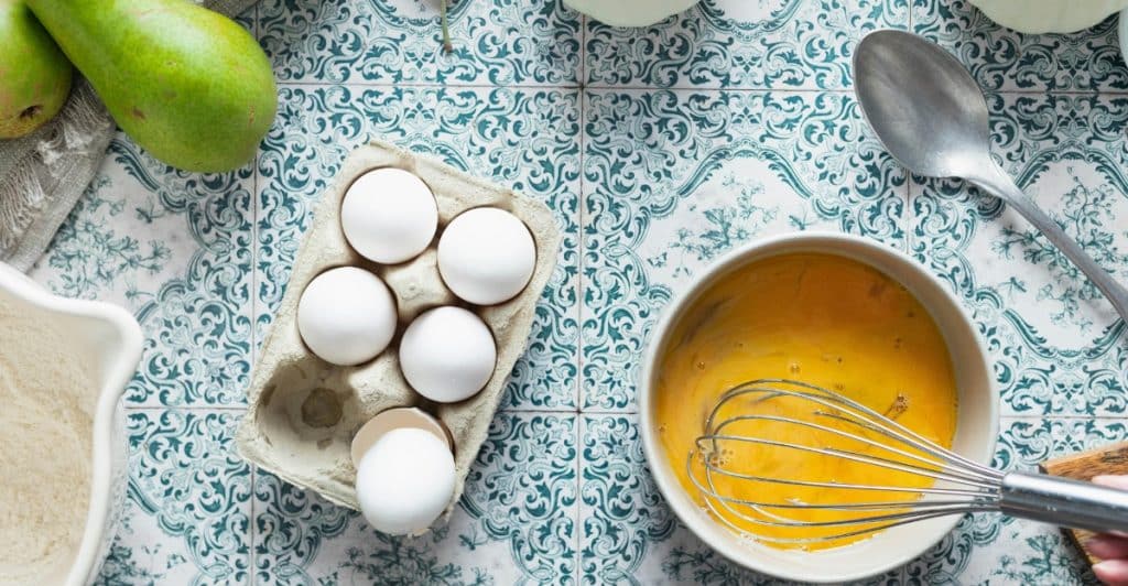 A carton of eggs and a bowl of beaten egg with a whisk on a patterned tile surface, with pears and other items in the background.