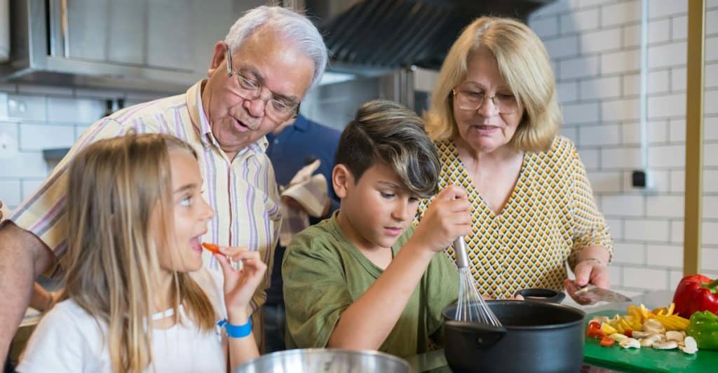 An elderly couple overseeing and helping their two young grandchildren, a girl and a boy, in the kitchen as they prepare food in a pot and bowl.