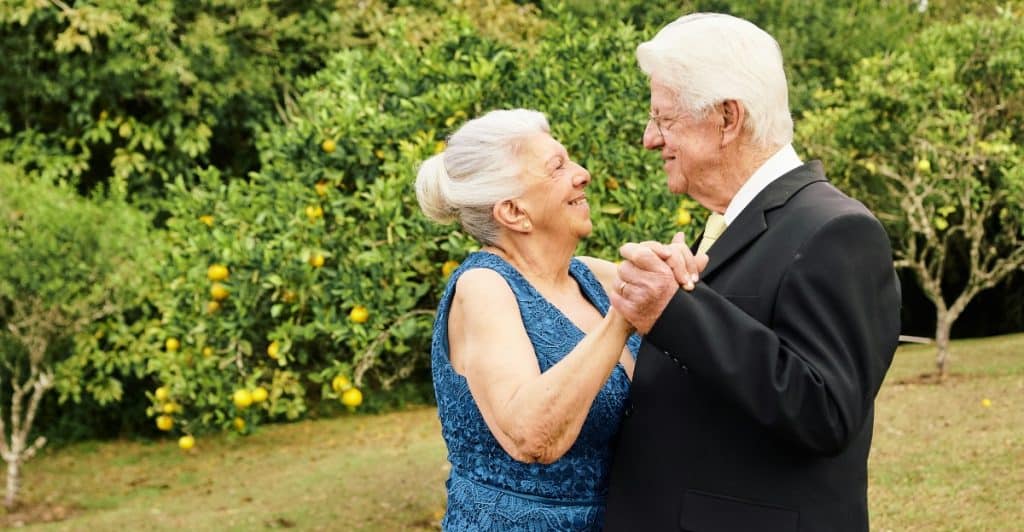 An elderly man in a suit and an elderly woman in a blue dress are dancing outdoors, holding hands and smiling at each other.