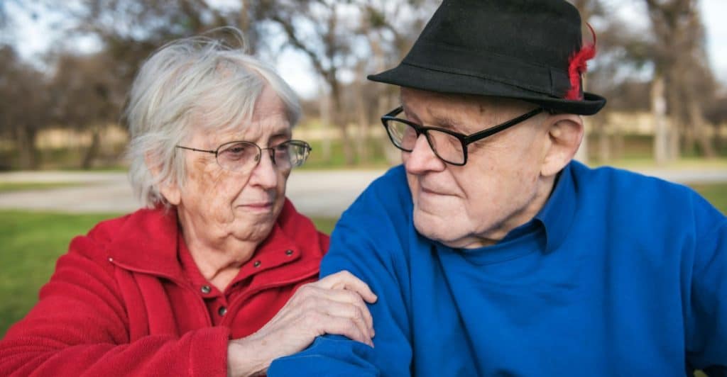 An elderly woman in a red jacket is sitting next to an elderly man in a blue shirt and a hat, and she is holding his arm.