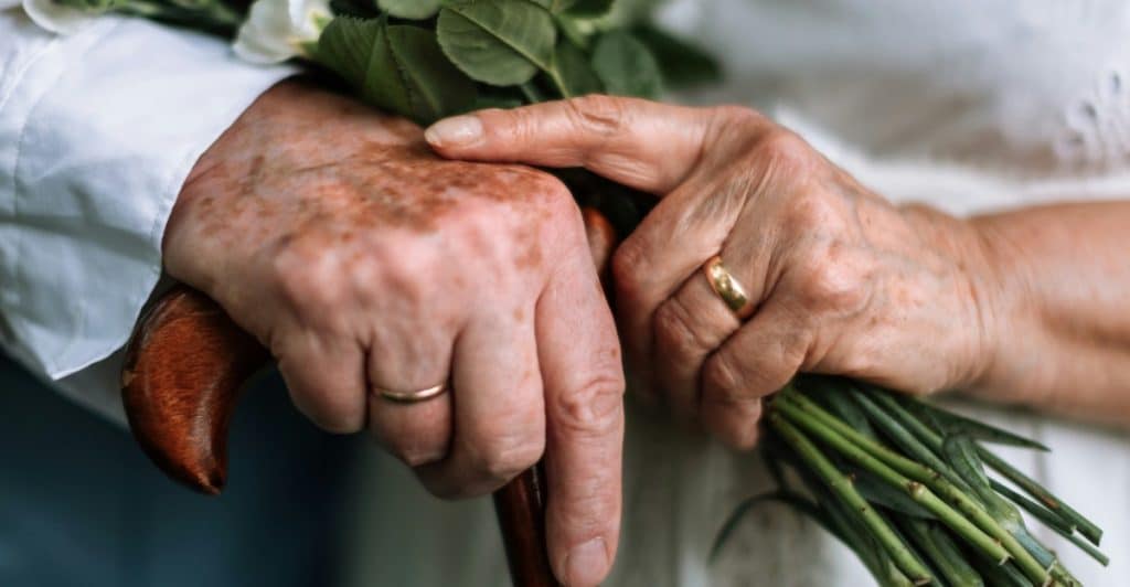 Close-up of elderly hands, one holding a walking cane and the other holding a bouquet of white flowers with green leaves