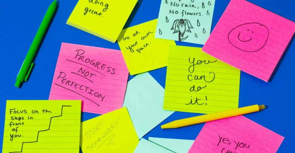 A collection of colorful sticky notes in neon green, pink, and yellow with various uplifting messages like "you're doing great" and "PROGRESS NOT PERFECTION".
