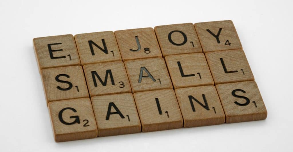 Wooden scrabble tiles are arranged to spell "ENJOY SMALL GAINS" on a white surface.