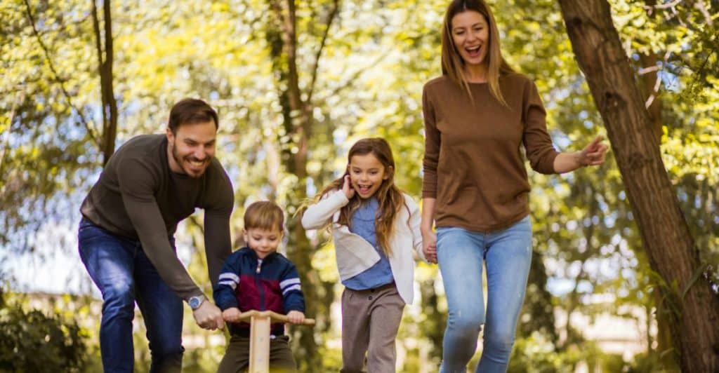 A family of four, including two adults and two young children, walking and smiling outdoors in a park.