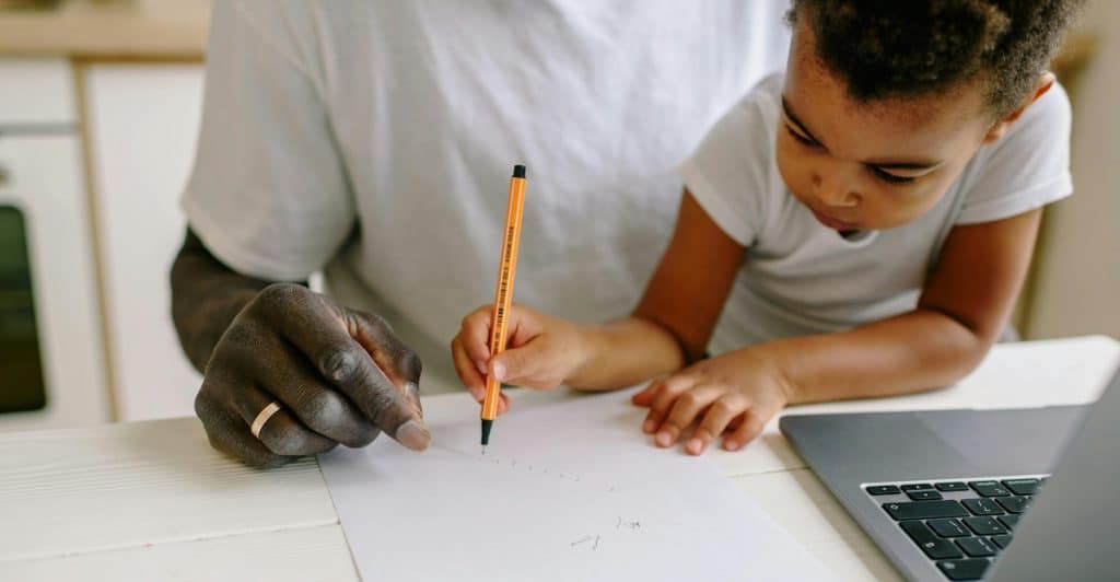 A man with a wedding ring on his left hand is seated at a light wooden table, helping a young child, possibly his son, draw on a white paper with a pencil, with a laptop open on the table to their left.
