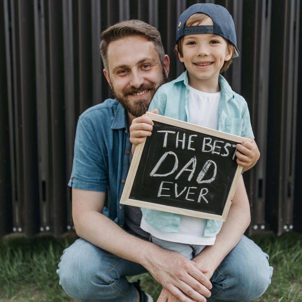 A father and son are crouching down, with the son holding a sign that says "THE BEST DAD EVER"