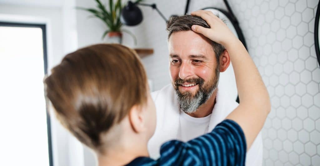 A man with a beard and a white towel over his shoulder is smiling and looking at a young boy who is facing away from the camera, with the man's hand touching the boy's head in a bathroom with hexagonal wall tiles.