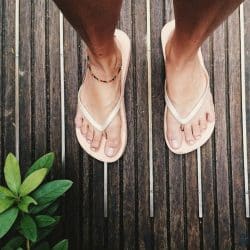 A top-down view of a person's feet wearing light-colored flip-flops, standing on a weathered wooden plank surface, with green leaves visible in the corner.