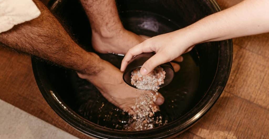 People's feet soaking in a dark basin while another person pours a grainy substance from a small bowl.
