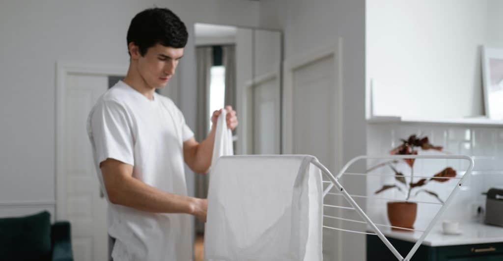 A man in a white t-shirt carefully folding a white sheet on a clothes drying rack in a well-lit room.