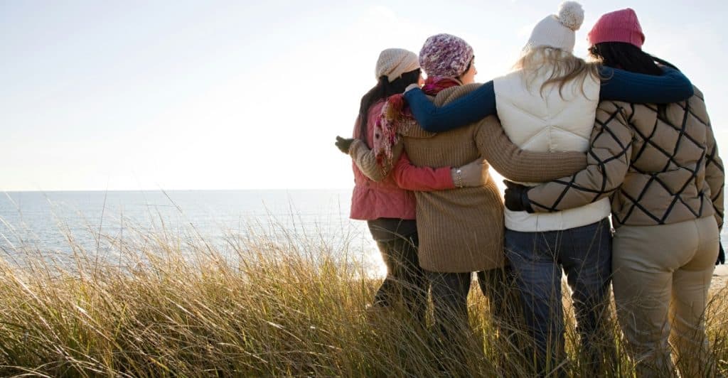 Four women with their arms around each other, looking out at the ocean from a grassy dune