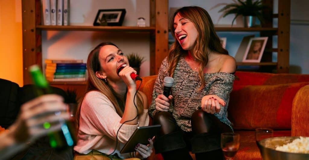 Two women singing karaoke on a couch with friends.