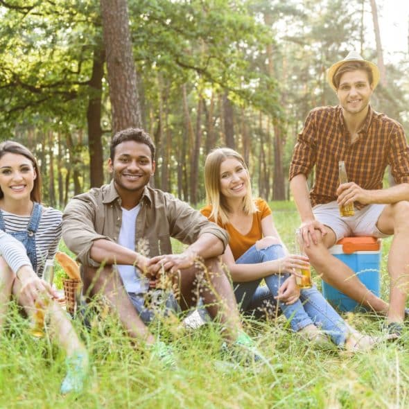 Four friends sitting on a blanket in a grassy, wooded area, smiling and looking at the camera.