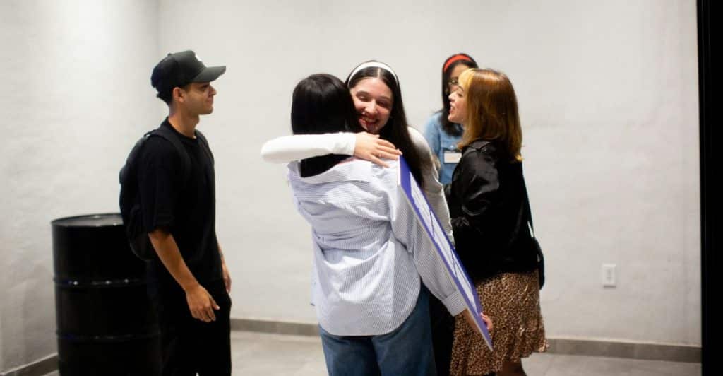 Two women embracing in a hug while other people stand in the background, suggesting a social gathering
