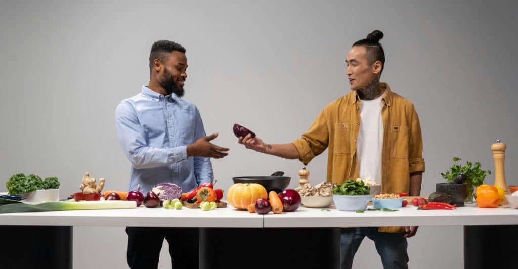 Two men of diverse backgrounds standing behind a table full of fruits and vegetable, with one man handing a piece of fruit to the other.