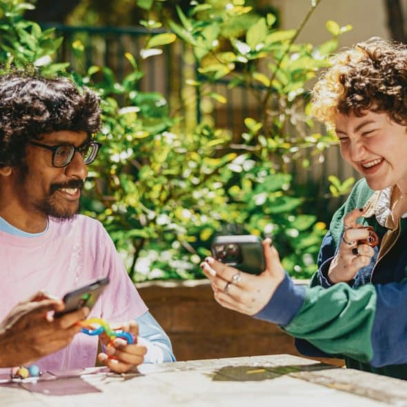 Two friends sitting at a table outdoors, looking at a smartphone and laughing.