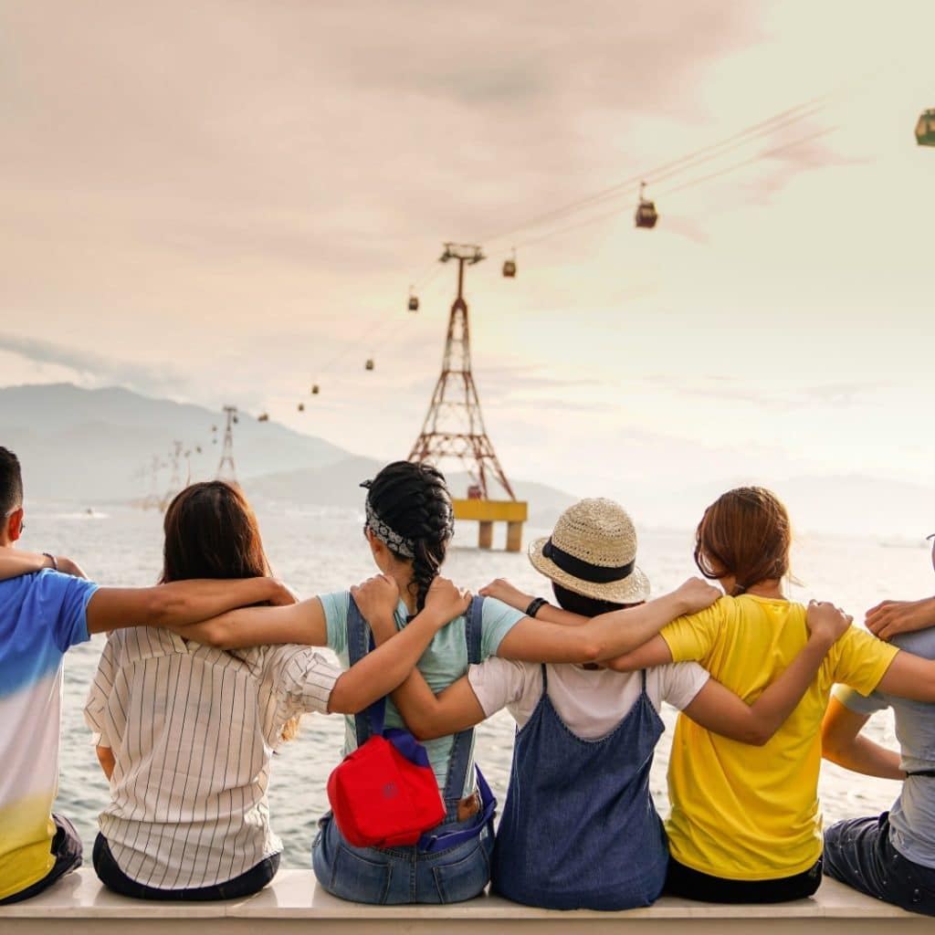 A diverse group of friends with their arms around each other's shoulders, sitting with their backs to the camera, looking out at a sunset over water with cable cars in the distance