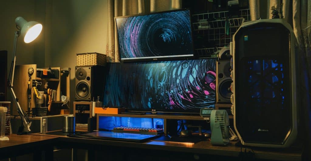 A gaming PC setup featuring a desktop computer tower with blue internal lights, multiple monitors displaying colorful abstract art, and large speakers.