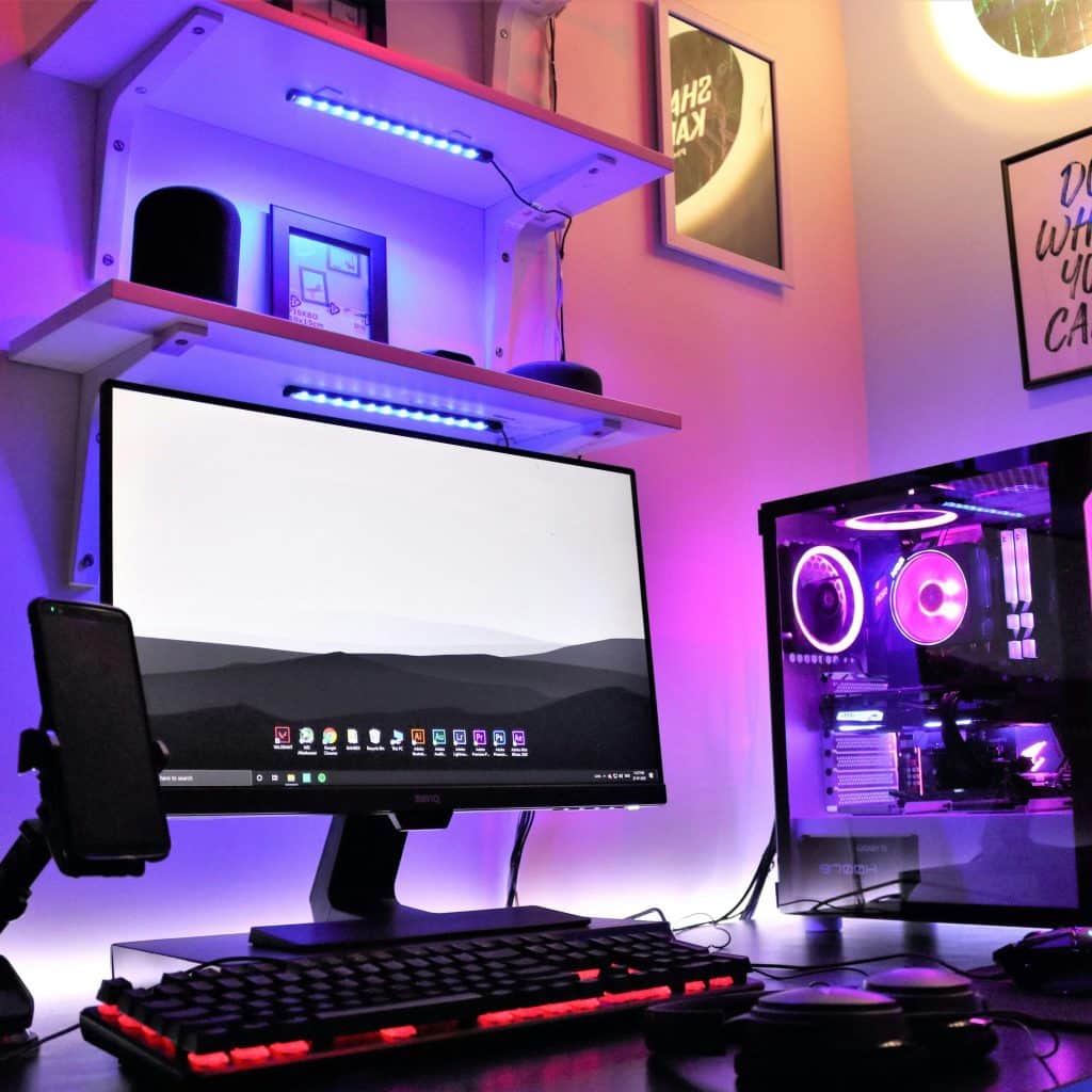 A gaming PC setup features a monitor displaying a monochrome landscape, a desktop tower with purple and blue internal lighting, and shelves with synchronized LED strips.
