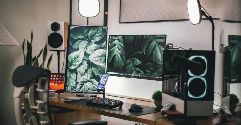 A gaming desk setup with two monitors, white bookshelf speakers, a tablet, and a desktop PC, adorned with green plant accents.