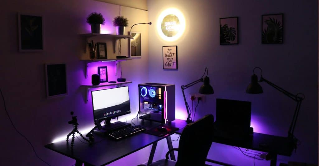 A gaming room illuminated with purple and white smart lighting, showcasing a desk with a monitor, a desktop PC, and ambient wall decor.