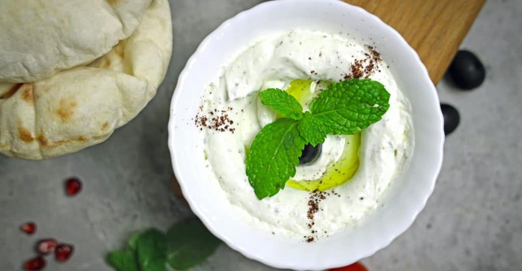 A white bowl holds creamy Greek yogurt garnished with green mint leaves and black olives, sprinkled with a dark spice, set next to flatbread, cherry tomatoes, and pomegranate seeds on a gray surface.