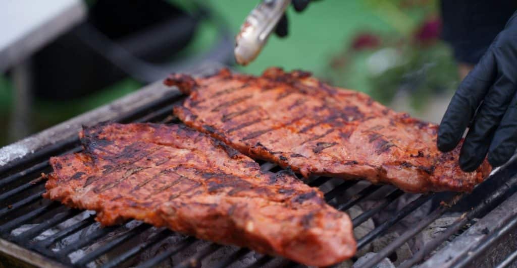Hands in black gloves using tongs to turn two large, seasoned cuts of meat with grill marks on an outdoor barbecue grate, with smoke rising.