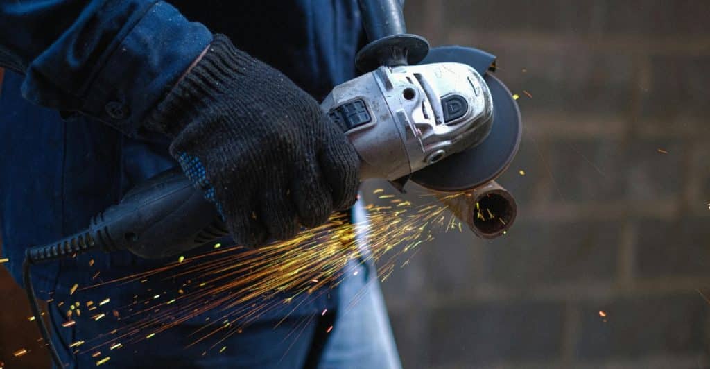 Close-up of gloved hands holding an angle grinder, with bright orange sparks flying off a piece of metal as it is being worked on.