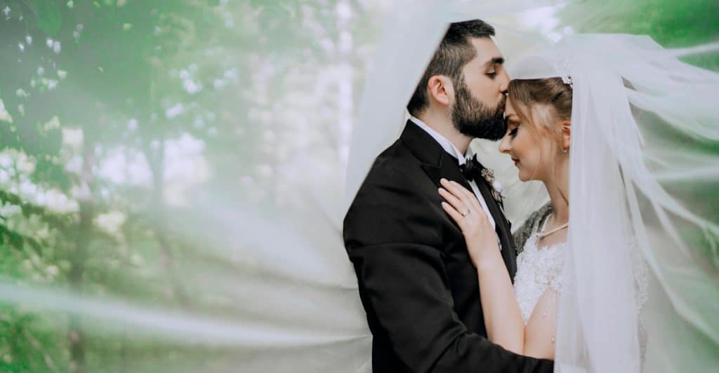 A groom kissing the forehead of his bride, both under a veil, in an outdoor setting with greenery.