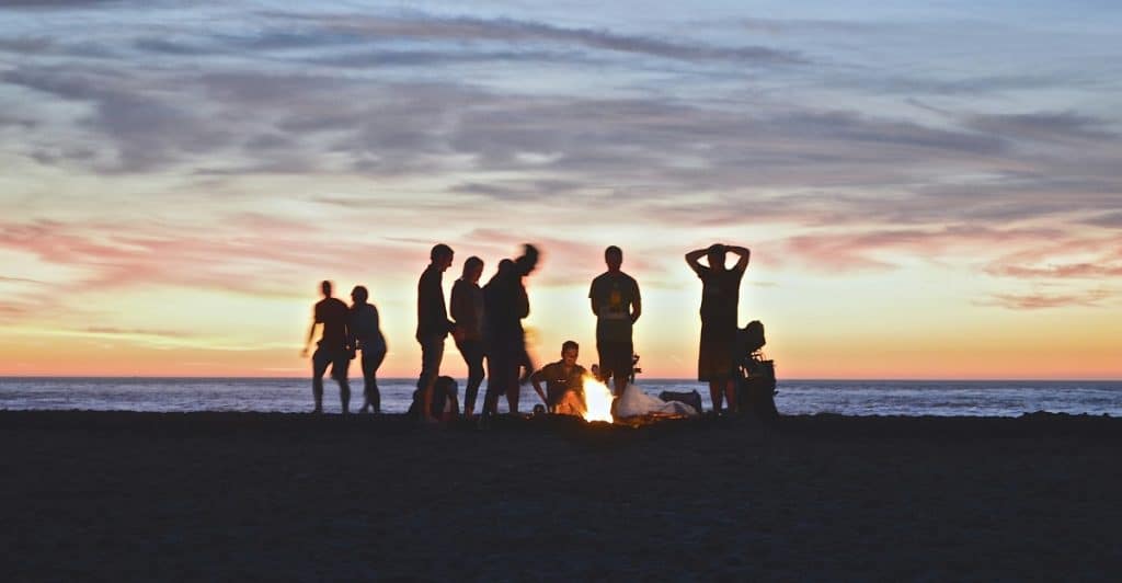 A group of friends silhouetted against a sunset over the ocean, gathered around a small fire.