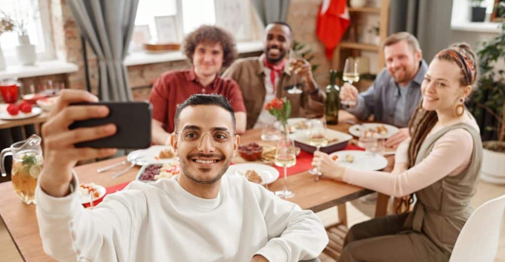 A diverse group of six friends is gathered around a long wooden dining table, with a man in the foreground smiling and holding up a phone to take a selfie of the group during a meal.