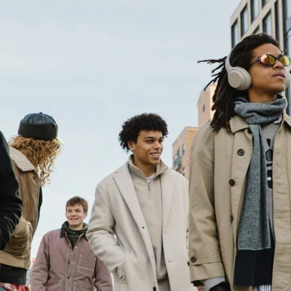 A group of men walking down a city street, one smiling in the foreground with headphones on and sunglasses.