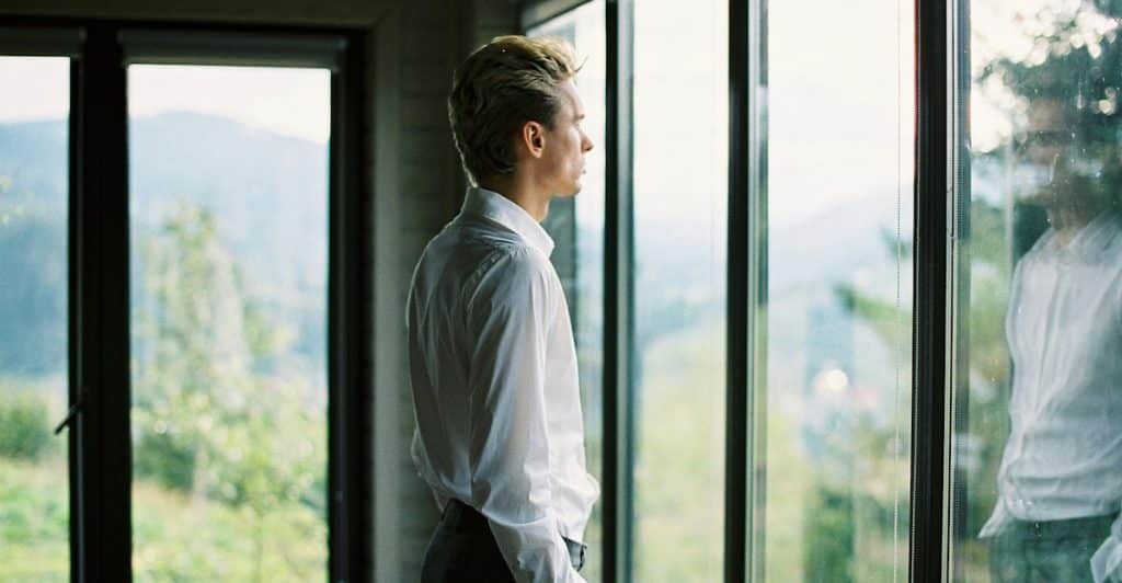 A man in a white shirt standing with hands in his pockets looking out a large window overlooking greenery.