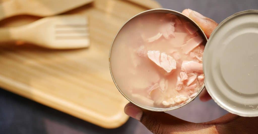 A person's hand holds an open, round tin can filled with flaked light pink tuna meat in water, with a wooden serving board and utensils blurred in the background.