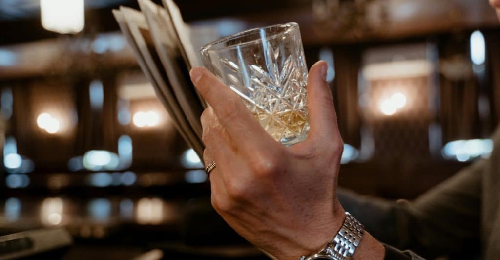 Close-up of a person's hand holding a glass of whiskey with a black dial watch on his wrist reading newspaper with his other hand.