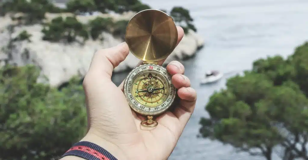 A person's hand is holding an open, gold-colored compass with a rotating bezel, overlooking a body of water and mountains