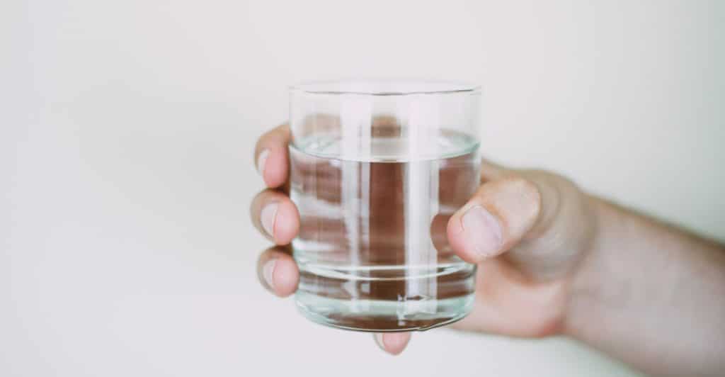 A hand holds a clear glass filled with water against a plain, light-colored background.