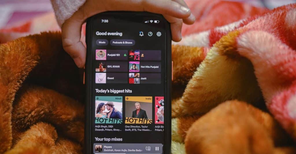 A person's hand holding a black smartphone displaying a music application with various playlists and song suggestions, resting on a patterned blanket.
