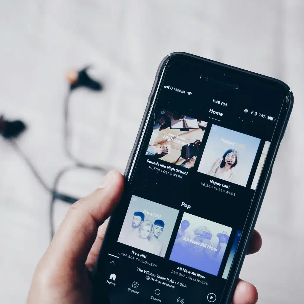 A hand holding a black smartphone displaying a music streaming application with various playlists and songs visible on the screen.