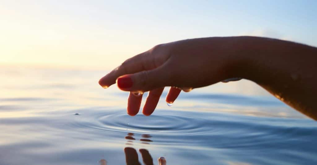 A hand with red nail polish gently touching the surface of water, creating ripples, with a bright sky in the background.