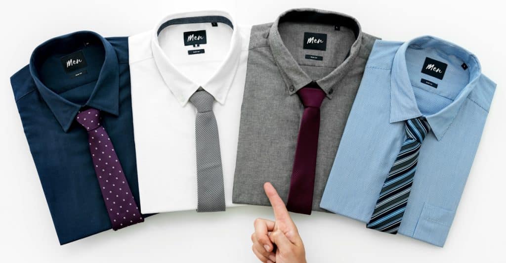 A hand with a gray suit sleeve is pointing towards four neatly folded men's dress shirts with ties, arranged side-by-side on a white surface.