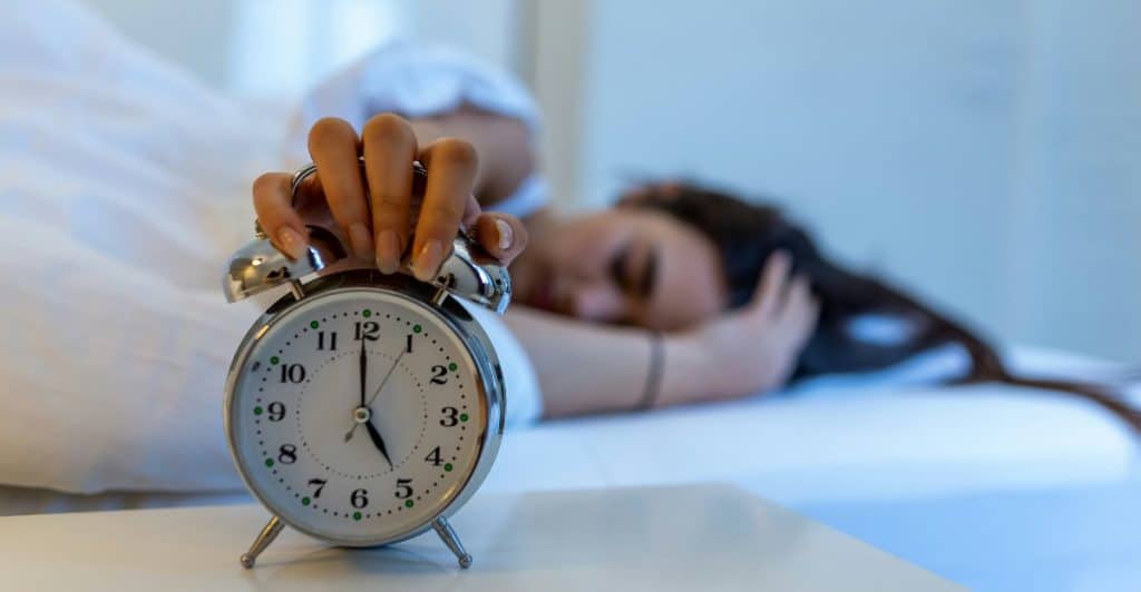 A hand reaching out to turn off an alarm clock next to a person sleeping in bed.