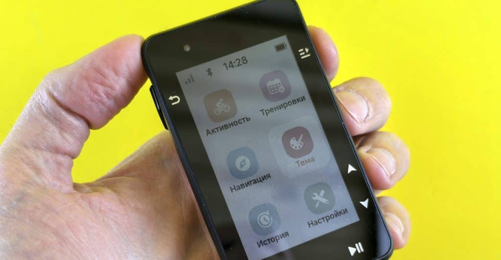 A hand holding a black handheld GPS device displaying various menu options like "Activity" and "Navigation" on its screen