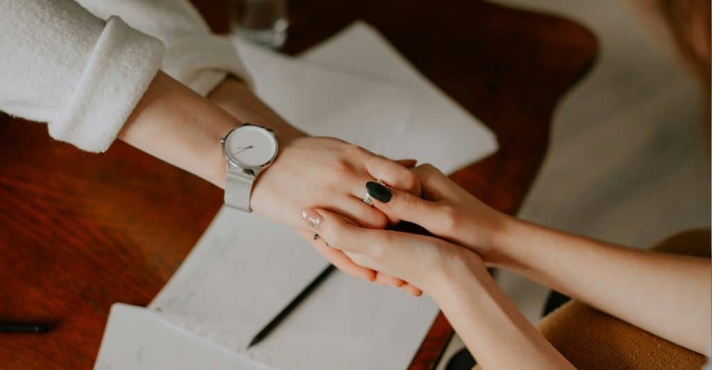 A close-up of two people's hands intertwined on a wooden table next to papers and a pen, one person wearing a watch.