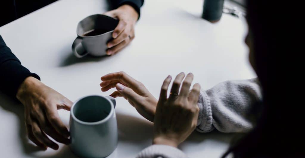 A high-angle view of two people's hands on a white table, with two gray mugs of coffee and one empty mug, suggesting a conversation.