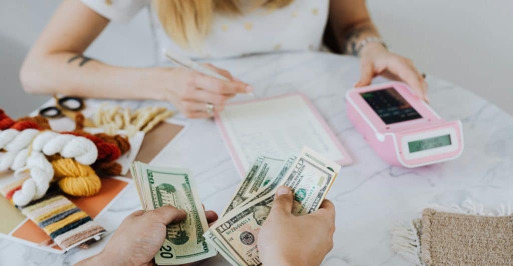 A person's hands holding cash over a table, with another person in the background writing and using a calculator, surrounded by craft supplies.